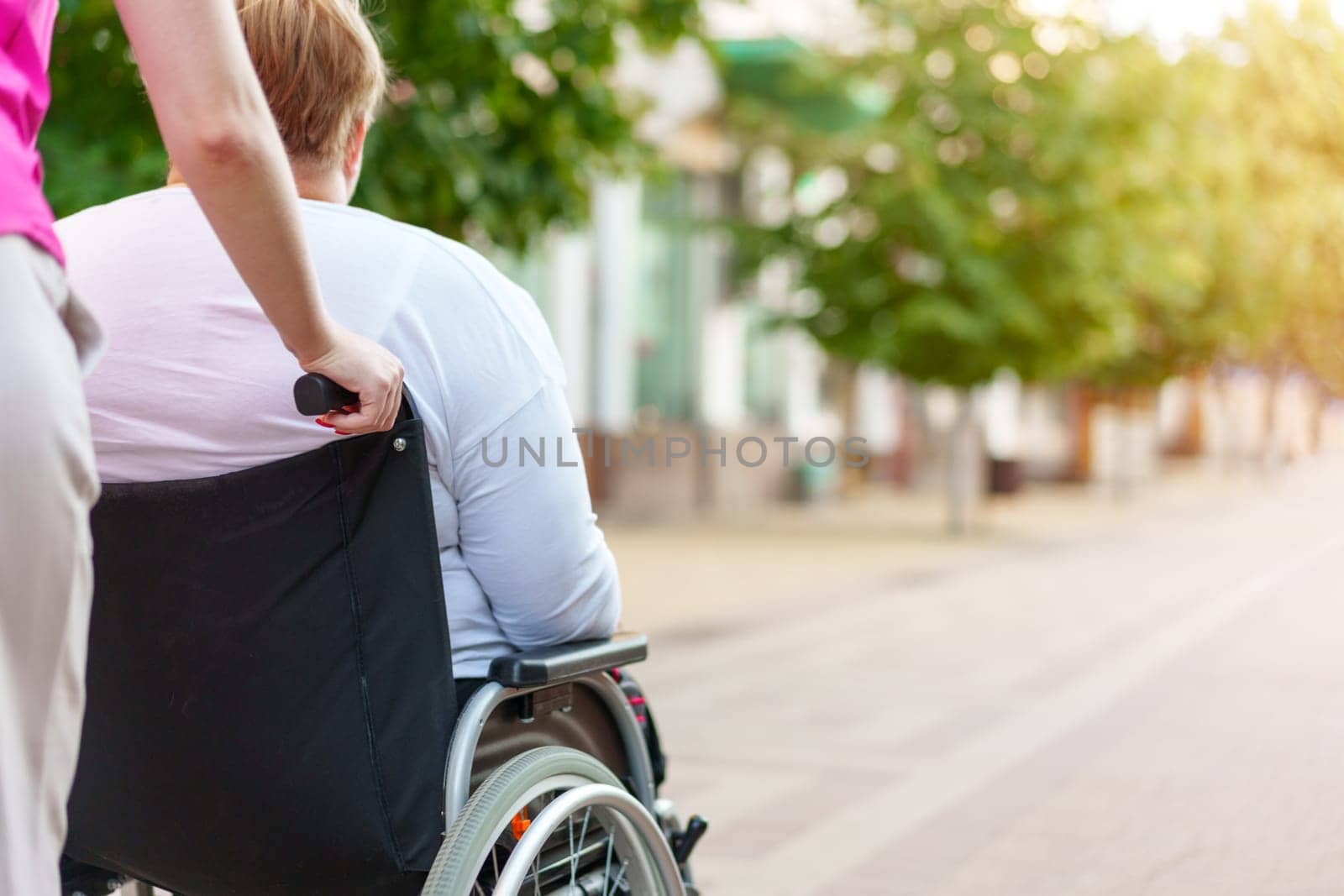 Back view of young woman helping mature woman in wheelchair strolling in the city
