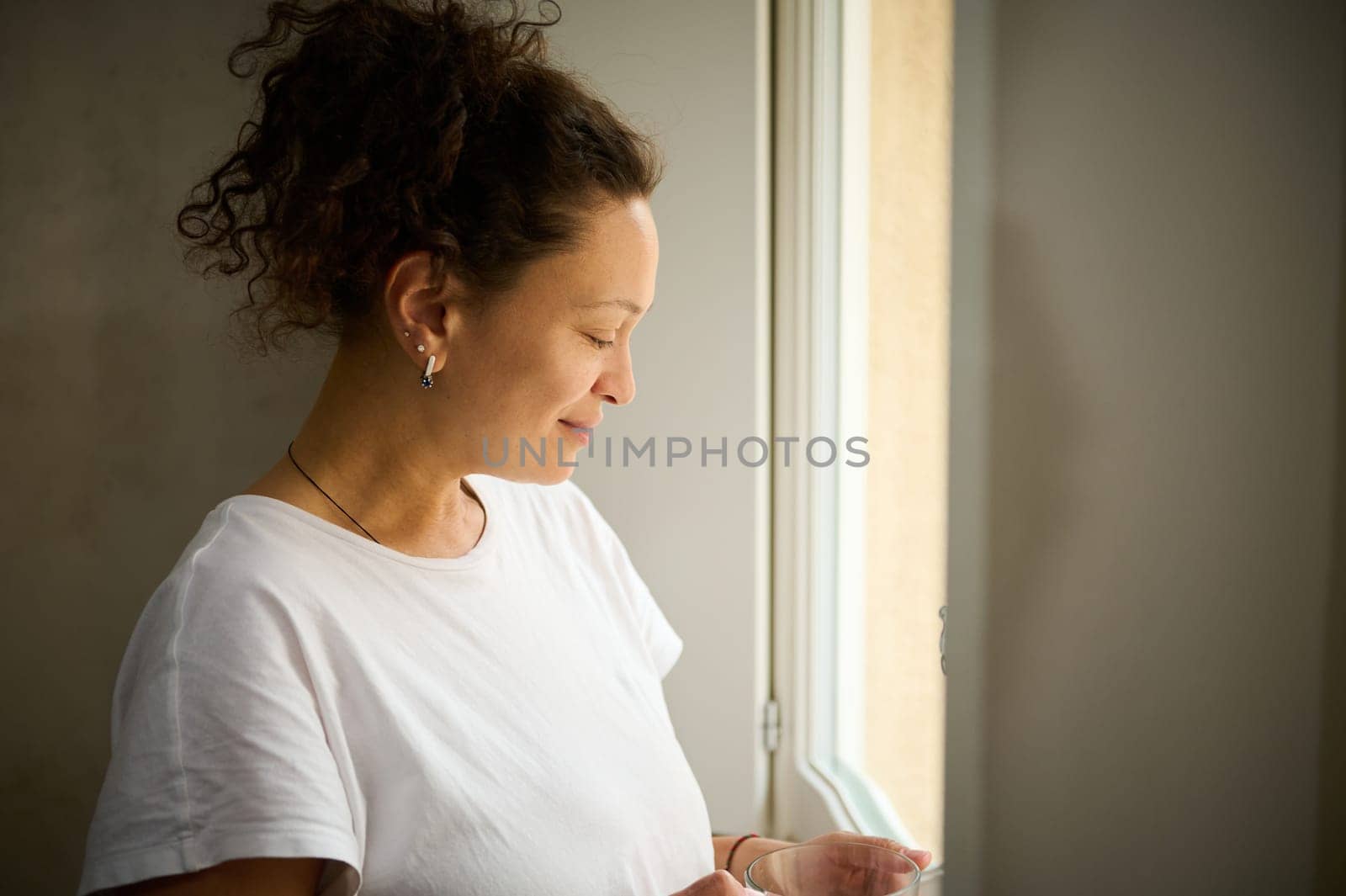 Close-up portrait of a multi ethnic curly haired woman holding a cup of hot drink, smiling admiring the view from her window by artgf