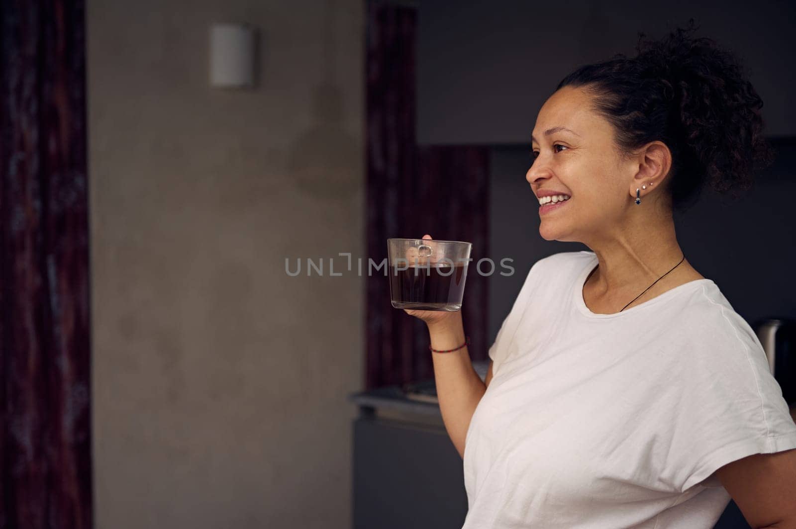 Multi ethnic curly haired pretty woman in white t-shirt of her pajamas, holding a cup of freshly brewed coffee drink, smiling looking away, enjoying her happy weekend at home. Copy advertising space by artgf