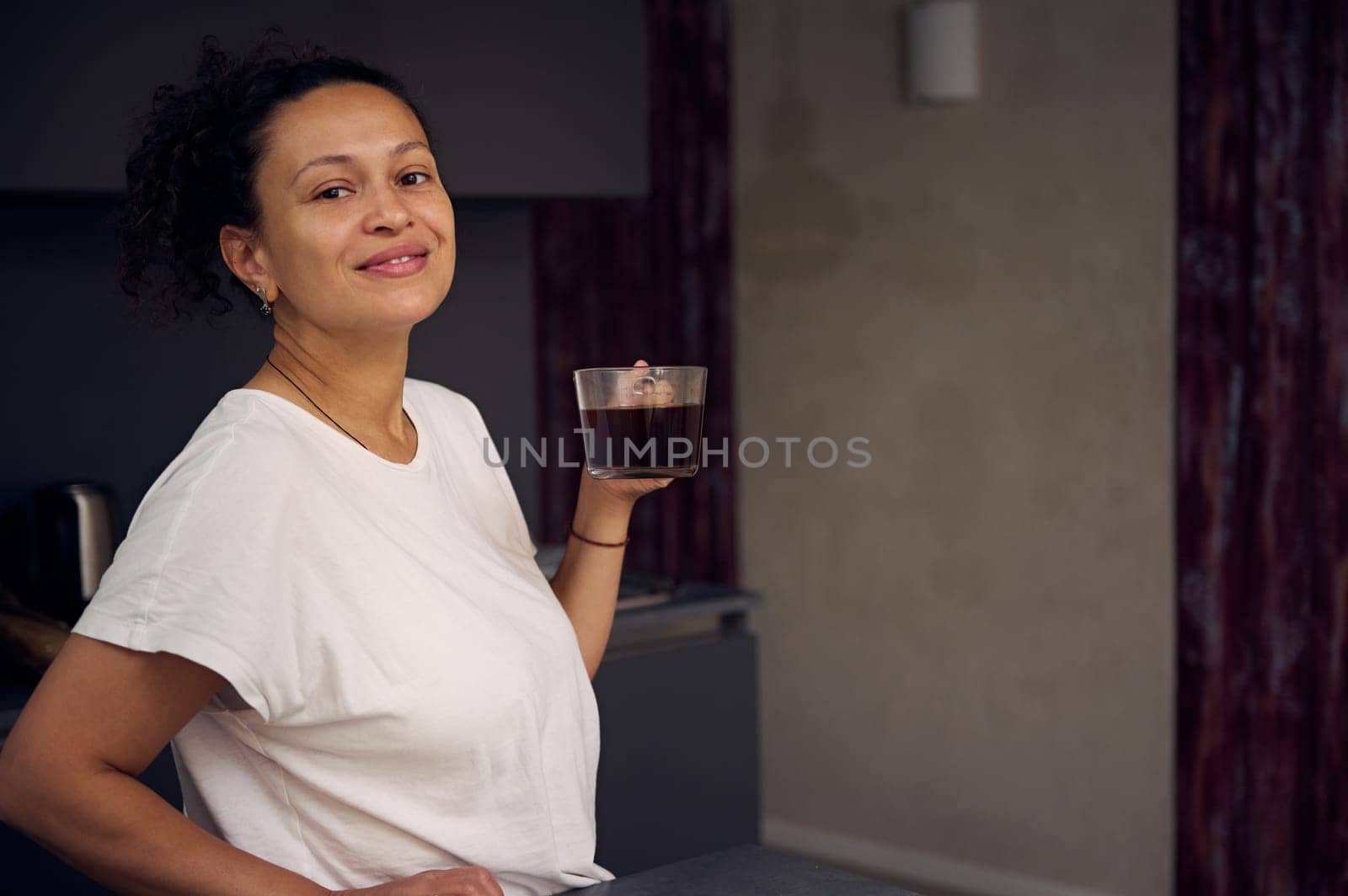 Young pretty woman smiling looking at camera while drinking coffee in the home kitchen. Start your day with energizing freshly brewed coffee. Copy advertising space