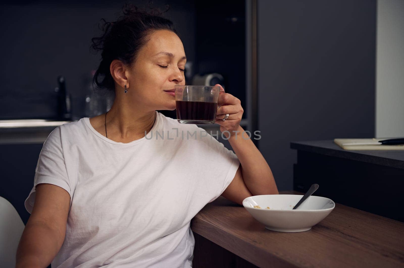Close-up portrait of a young pretty woman in white pajamas, sitting with her eyes closed at the kitchen table, taking sip of a freshly brewed espresso coffee. People. Domestic life. Hobbies and leisure