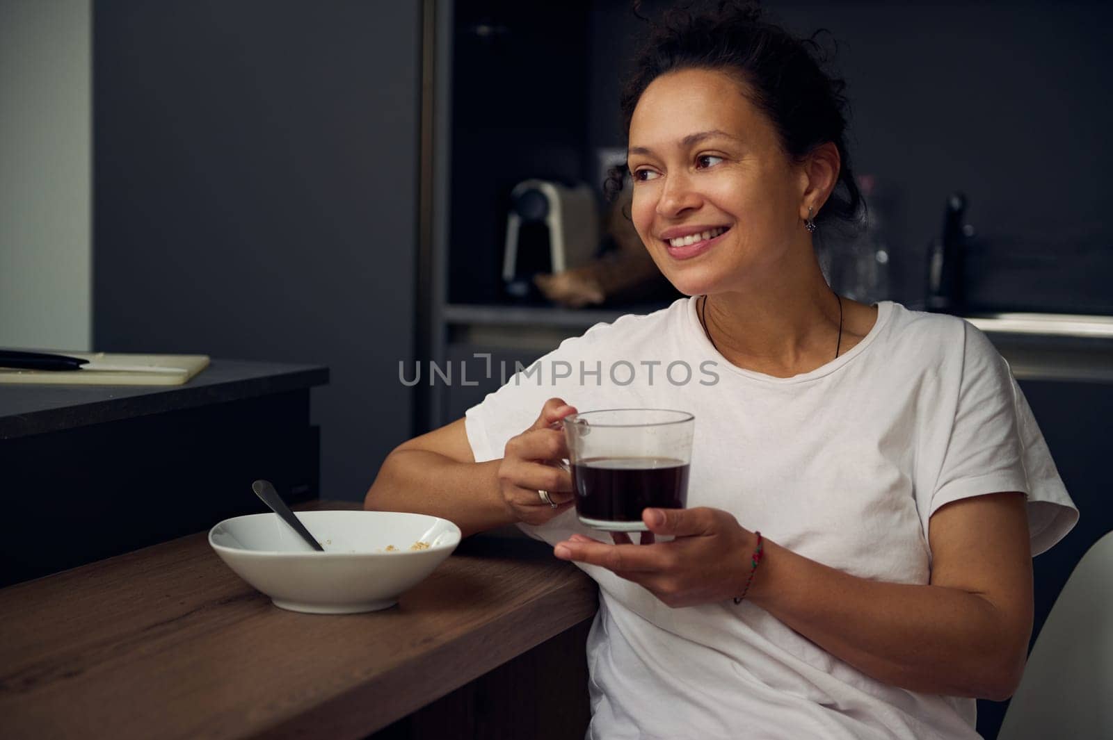 Portrait of a joyful young woman enjoying a cup of coffee at home. Attractive pretty lady drinking hot freshly brewed coffee, smiling looking away, sitting at kitchen table while taking a breakfast