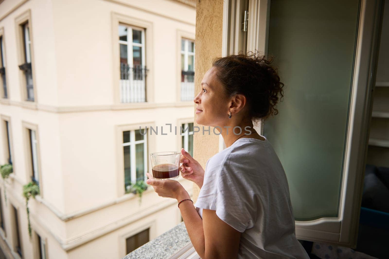 Happy woman admiring the view of a beautiful city overlooked from her house, standing at window with a cup of rom coffee in the sunny morning. Pretty tourist drinks coffee and admire the Como city