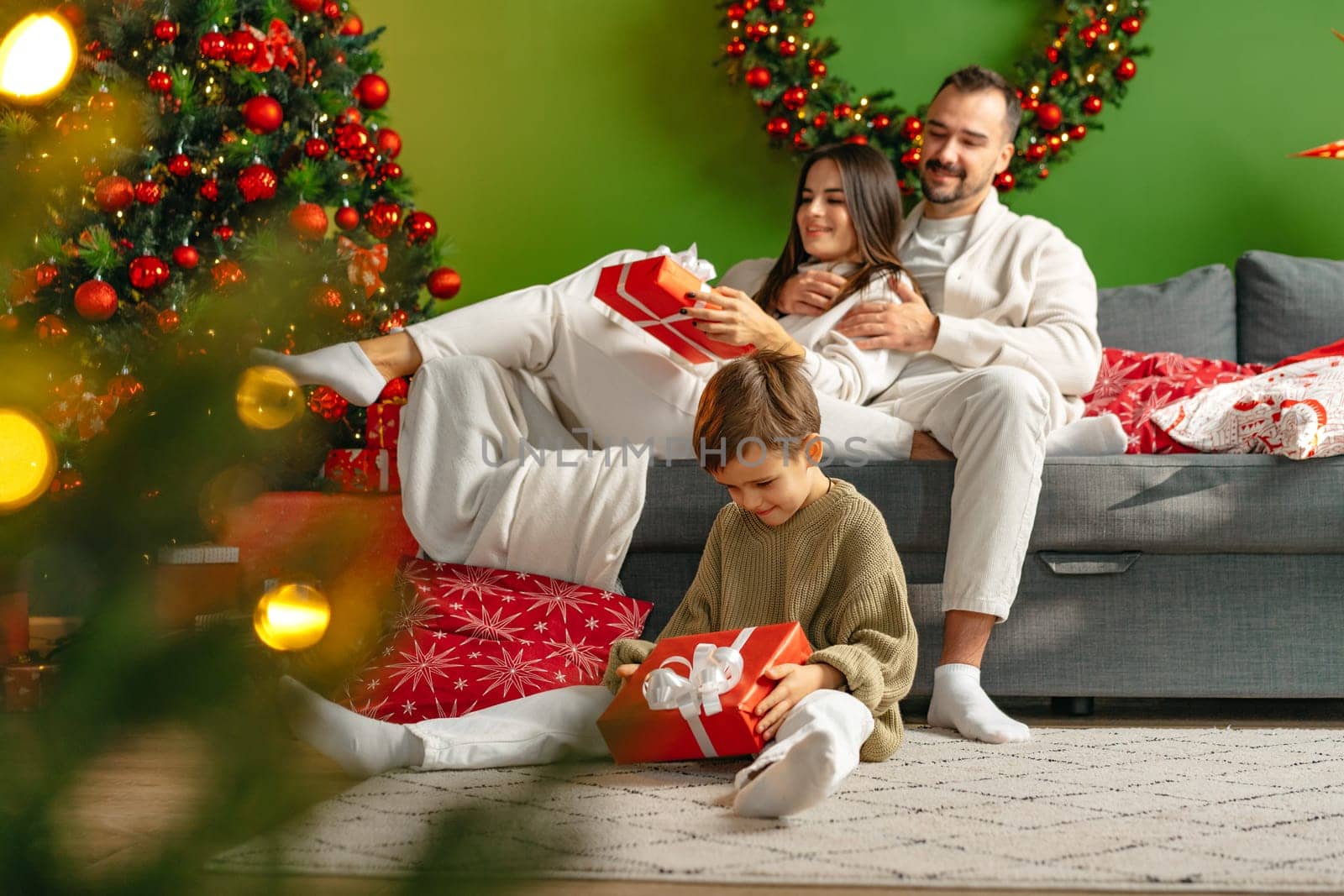 Happy young family sitting on sofa and relaxing at home at Christmas by Fabrikasimf