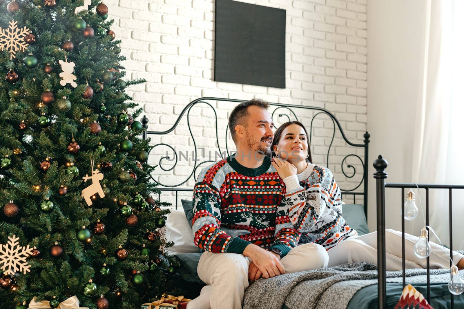 Young woman and man sitting on bed in sweaters and hugging near Christmas tree close up