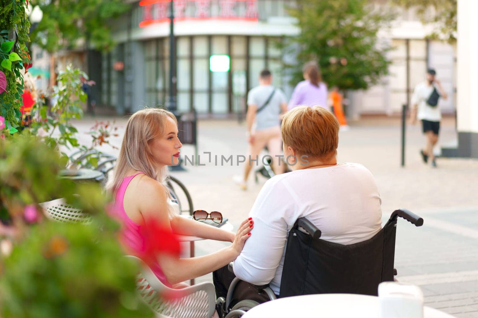 Woman with disability sitting in a wheelchair in outdoor cafe with her young daughter by Fabrikasimf