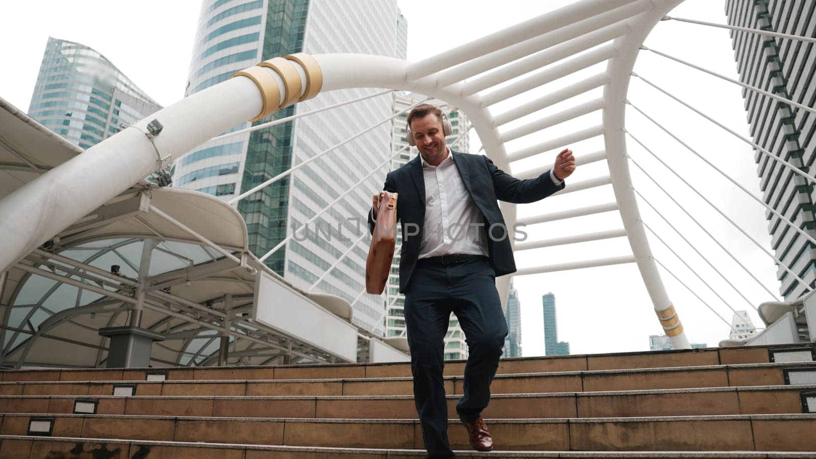 Happy businessman walking down stair and moving to music from headphone. Low angle camera view of smart manager listen song by using heaset and going back to home in urban city in lively mood. Urbane.