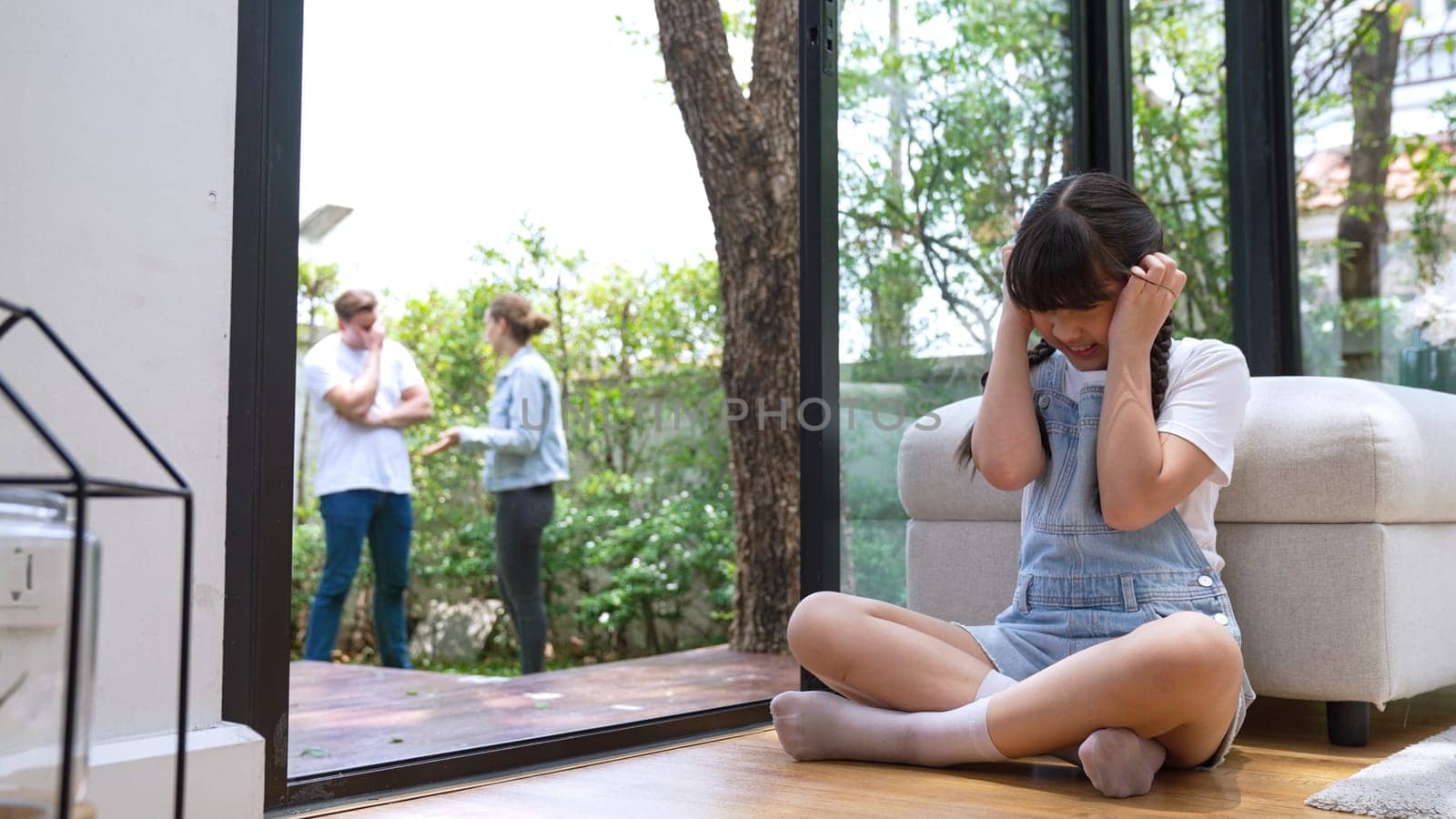 Stressed and unhappy young girl huddle in corner, cover her ears blocking sound of her parent arguing in background. Domestic violence at home and traumatic childhood develop to depression. Synchronos