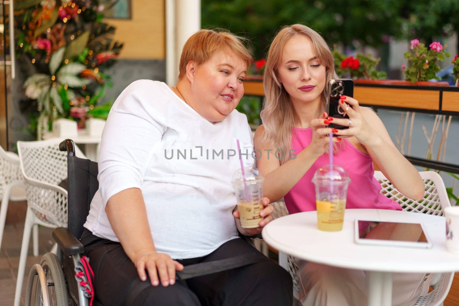 Portrait of mature mother in wheelchair and her daughter making a selfie using smart phone in cafe by Fabrikasimf