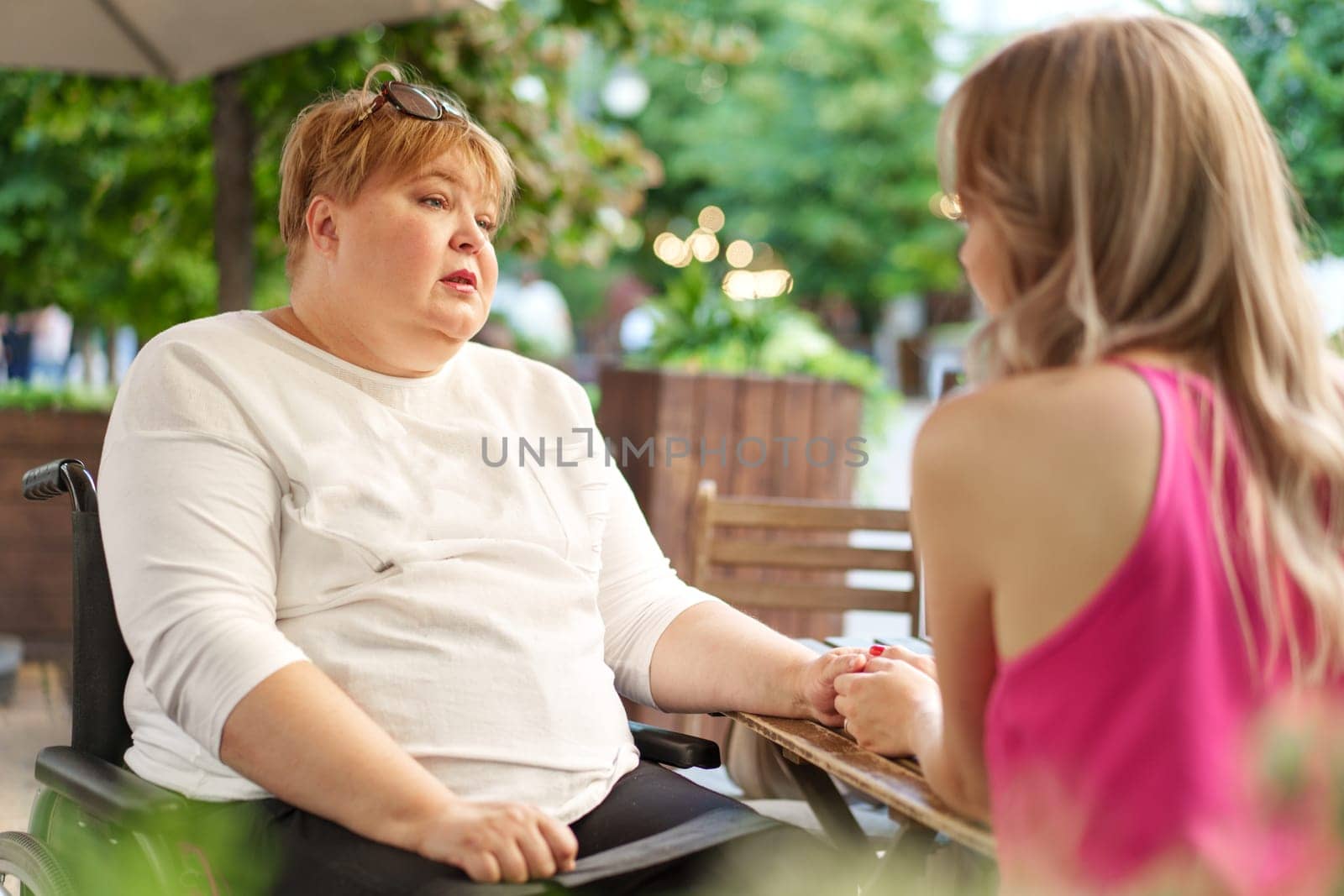 Mother with disability in wheelchair talking to her daughter while sitting at the table in cafe in the street