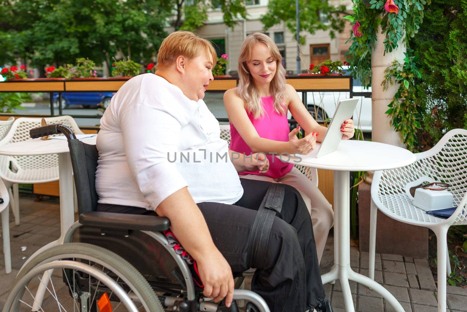 Mother with disability in wheelchair and her daughter using digital tablet while sitting at the table in cafe by Fabrikasimf