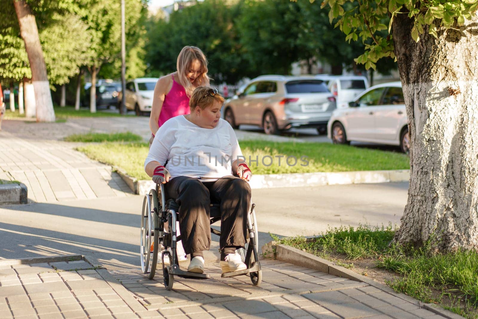 Young female caregiver pushing wheelchair with female person with disability across city street by Fabrikasimf