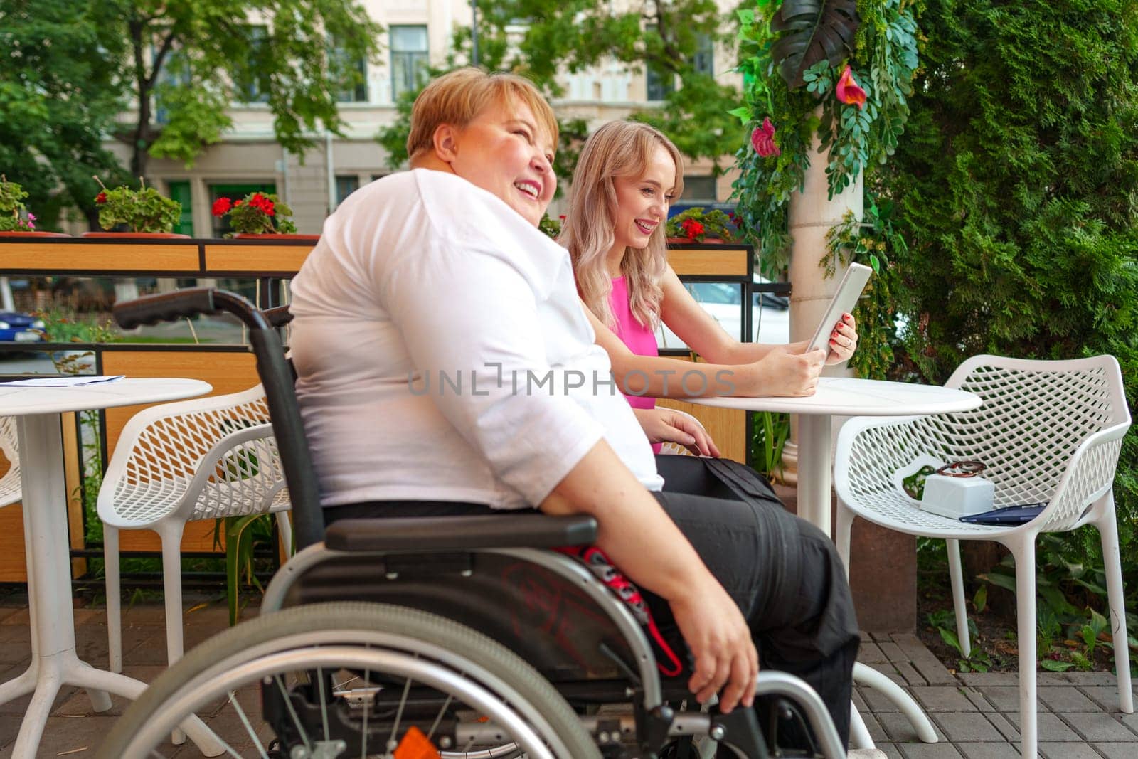 Mother with disability in wheelchair and her daughter using digital tablet while sitting at the table in cafe by Fabrikasimf