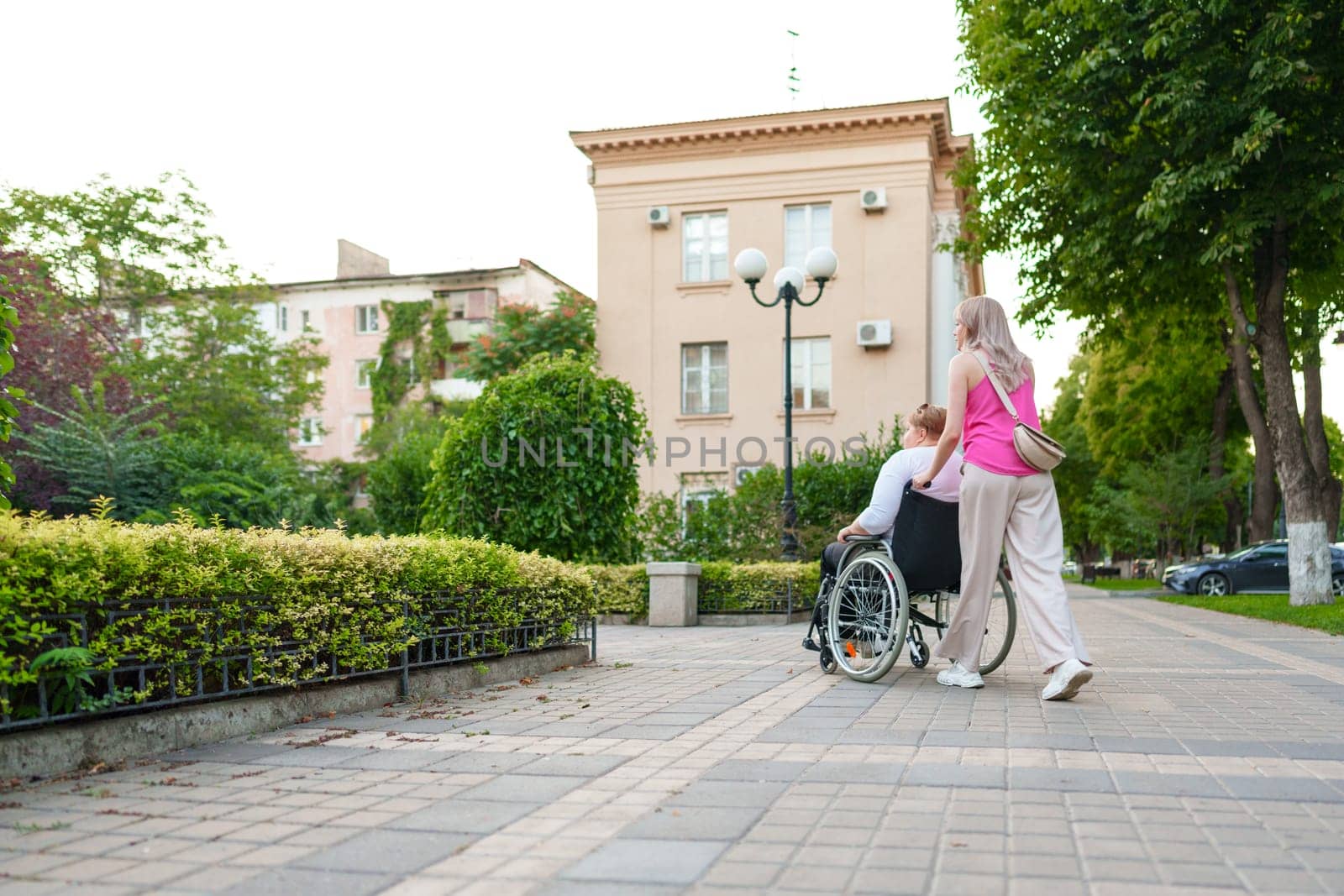 Back view of young woman helping mature woman in wheelchair in the city by Fabrikasimf