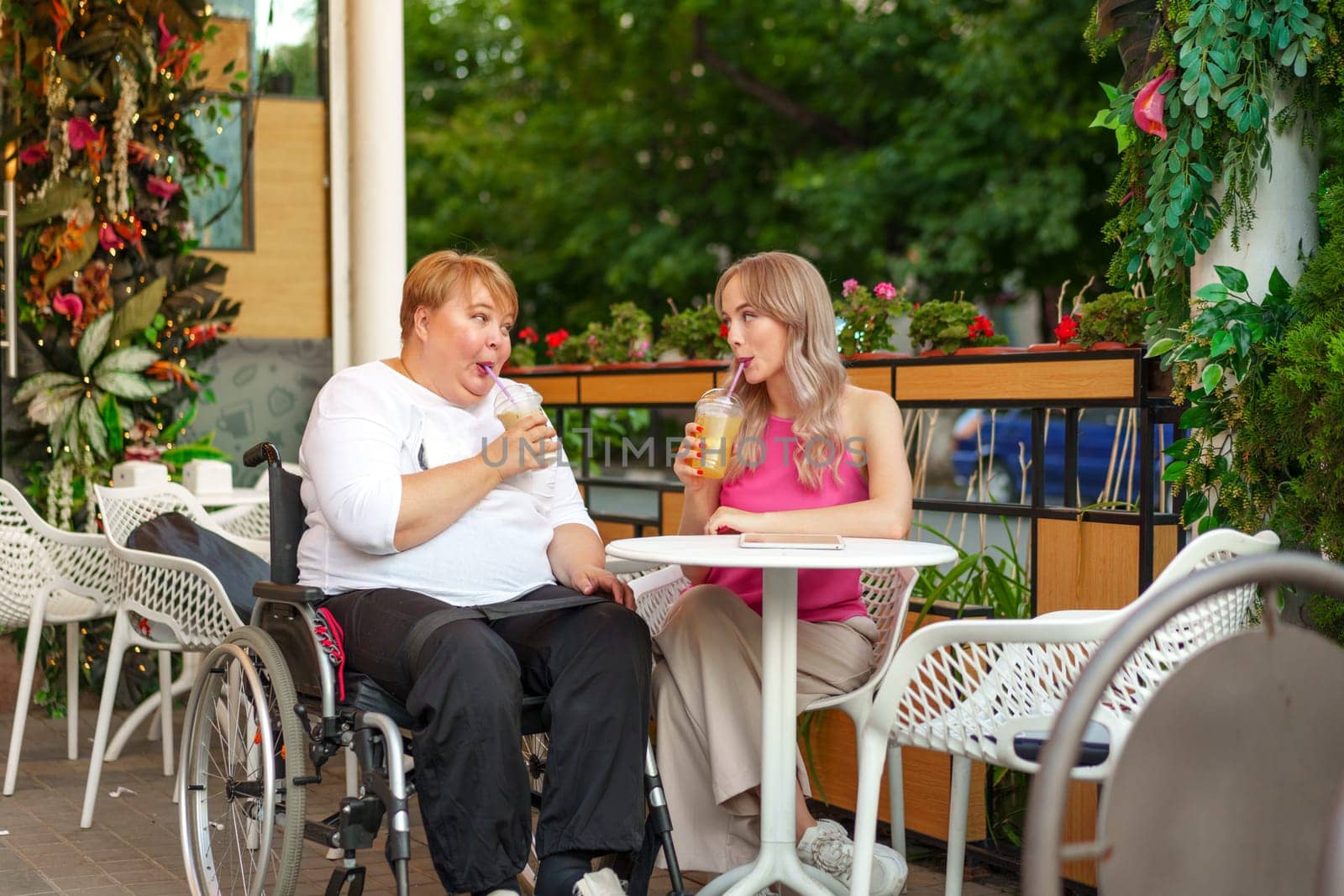 Young daughter and her mother in wheelchair sitting at the table in cafe with drinks and having fun by Fabrikasimf