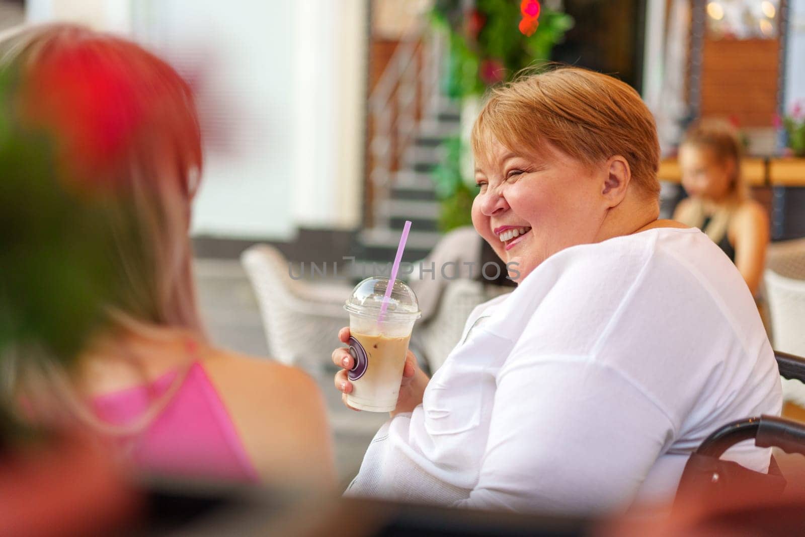 Young daughter and her mother in wheelchair sitting at the table in cafe with drinks and having fun, close up