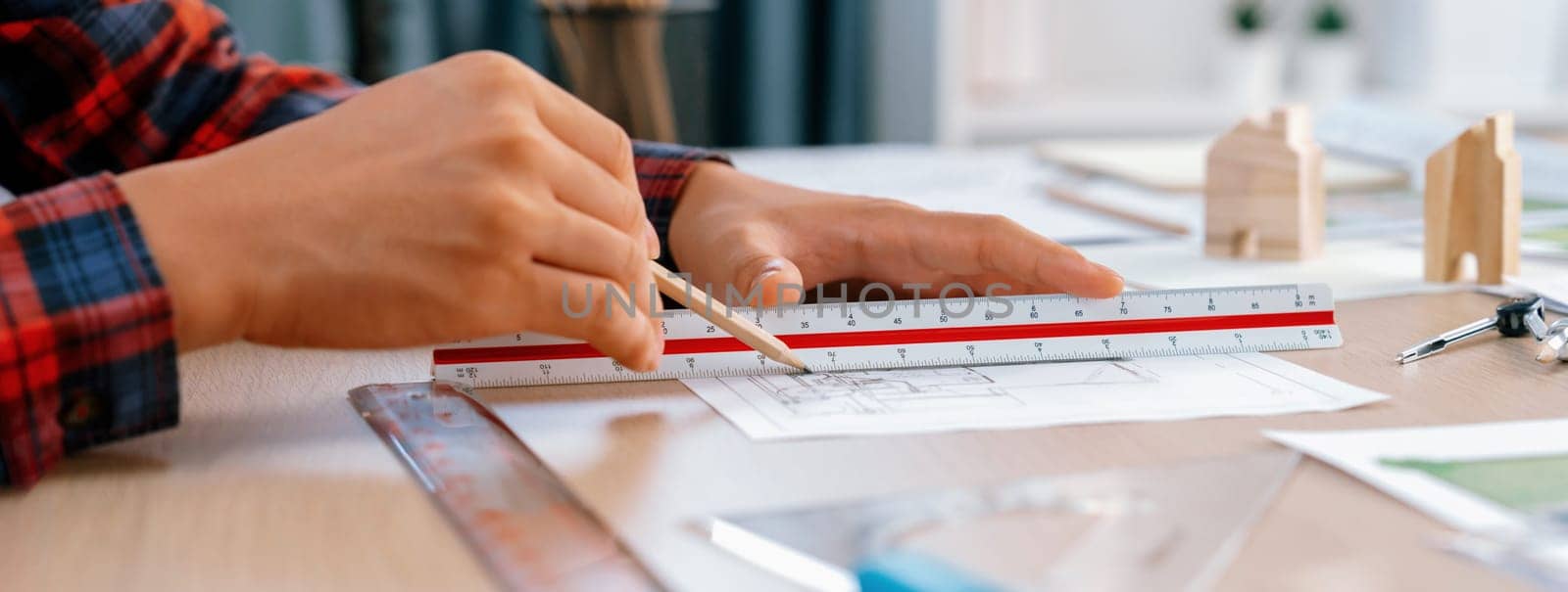 Closeup of architect engineer hand using ruler to mature and draw a blueprint on meeting table with wooden block, pencil and blueprint scatter around at architectural modern office. Delineation.