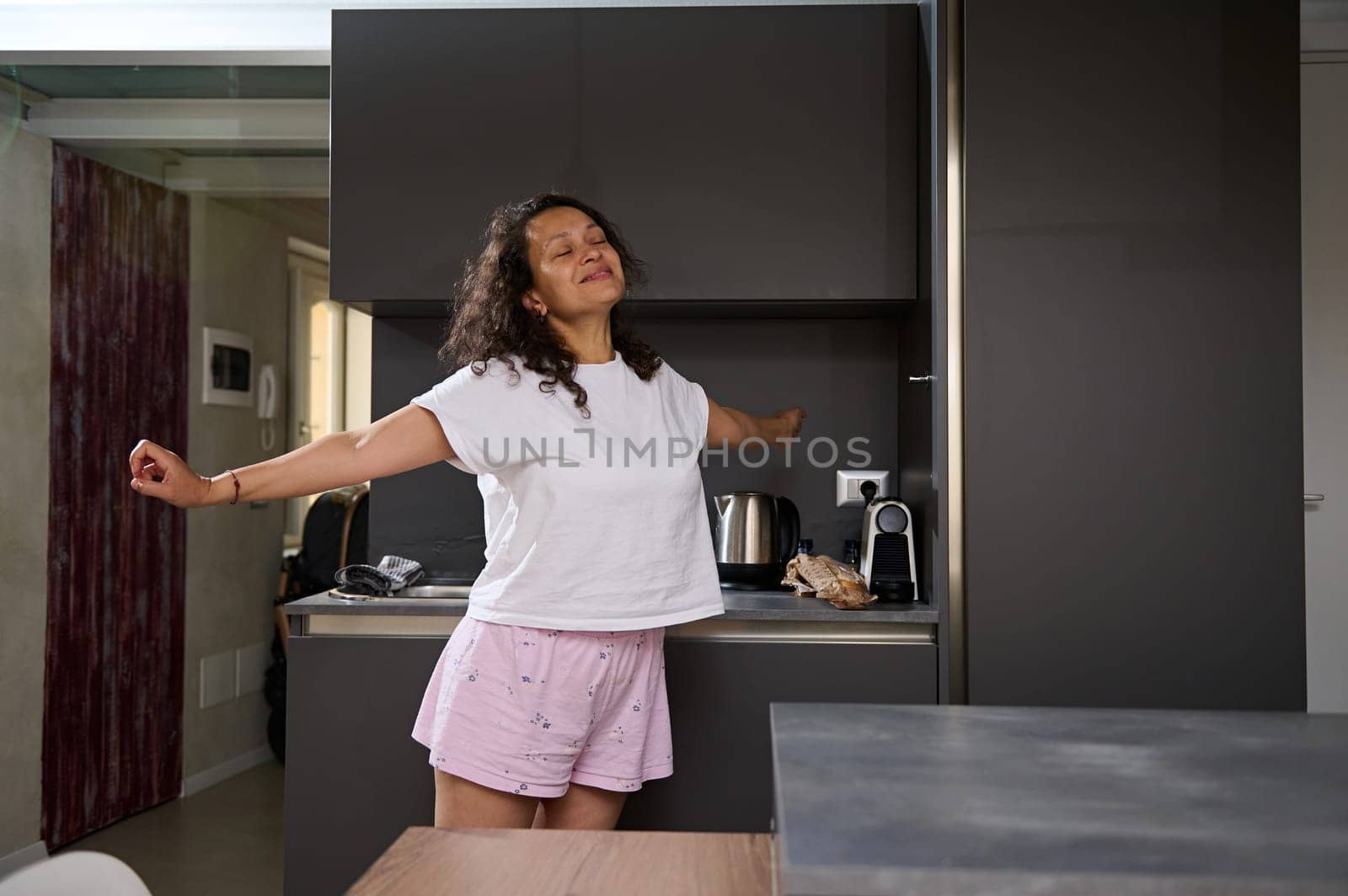 Portrait of a multi ethnic curly haired happy young woman in pajamas, stretching and smiling standing in minimalist modern kitchen. Cheerful female stretching and waking up early in morning at home