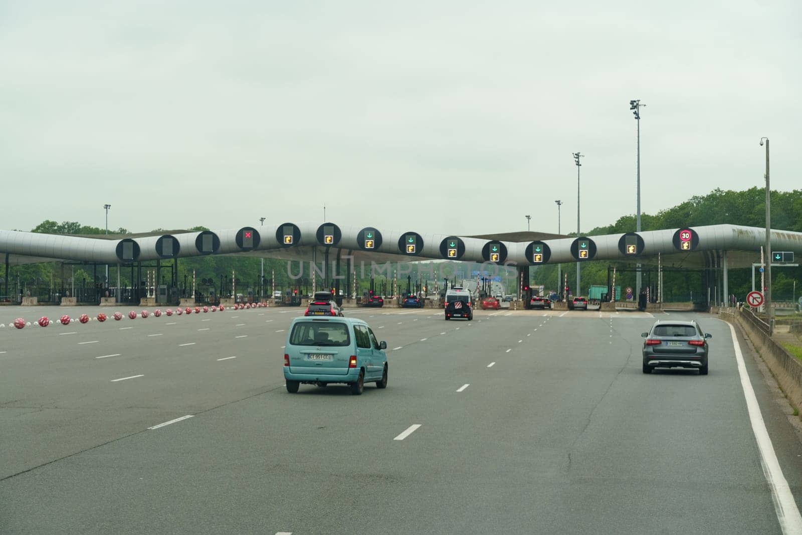 Lille, France - May 23, 2023: Vehicles line up to pay tolls at a modern toll plaza with multiple booths and electronic lane indicators on a cloudy day.