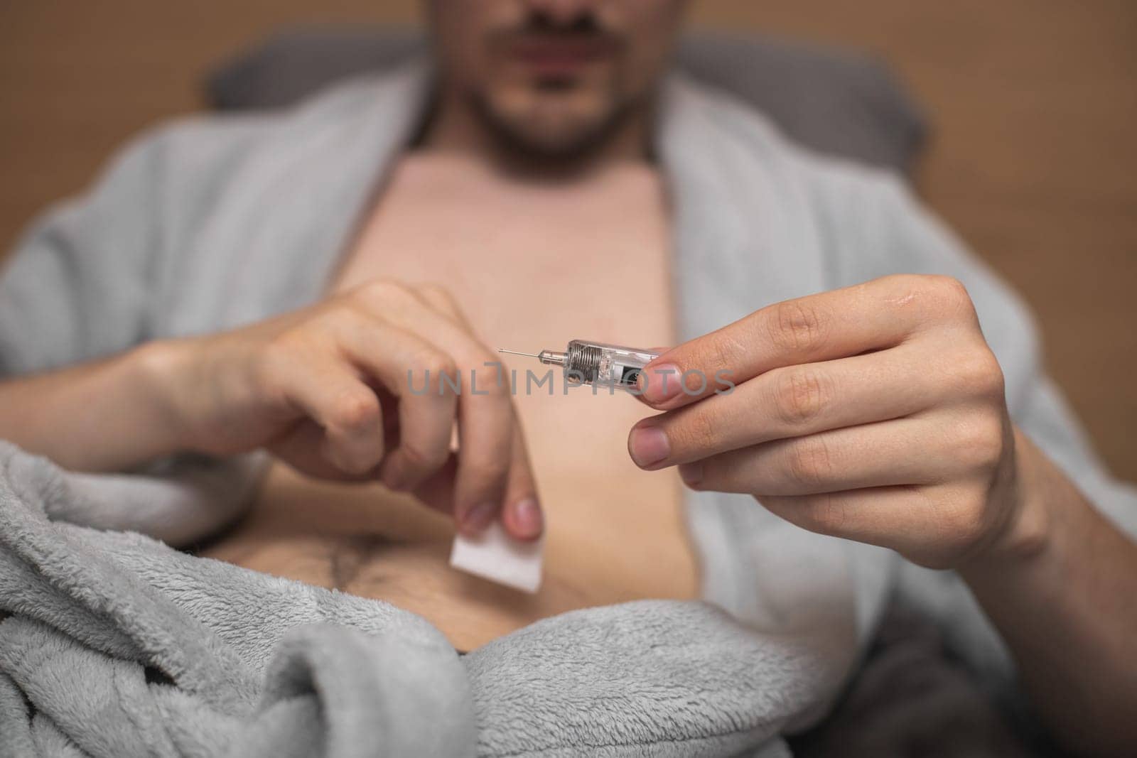 Step 4.Hands of a young caucasian man in a gray robe with a bared belly hold a syringe with a needle and medicinal liquid for an injection,lying on a bed,side view close-up.Concept step by step instructions for making an injection.