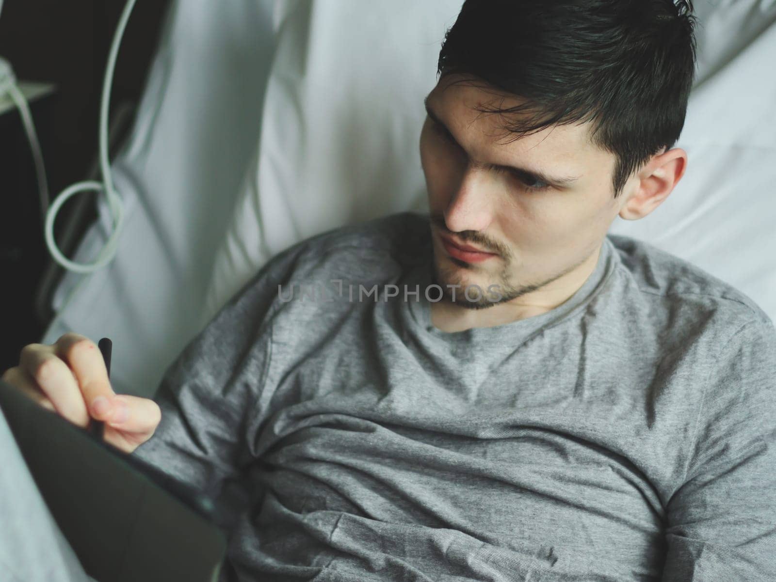 Young guy patient lying in bed holding a tablet talking with friends on social networks in the hospital, side view close-up with selective focus.Using technology and healthcare concept.