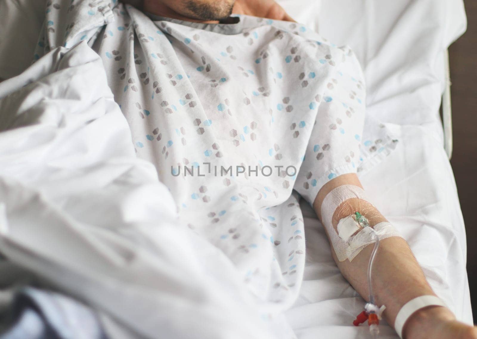The hand of a caucasian young male patient with a dropper in a vein attached with medical adhesive tape lies in bed with white bed linen and in a medical shirt, close-up top view. The concept of treatment of diseases, drug addiction.