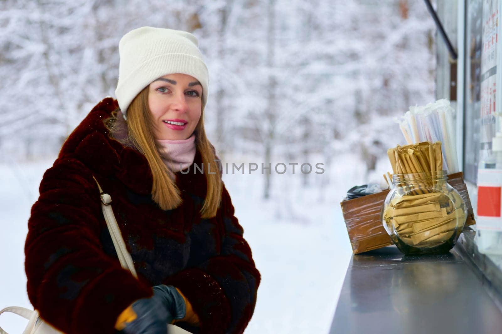 Pretty woman buying a hot drink at a kiosk in a winter park, snow covered trees by jovani68