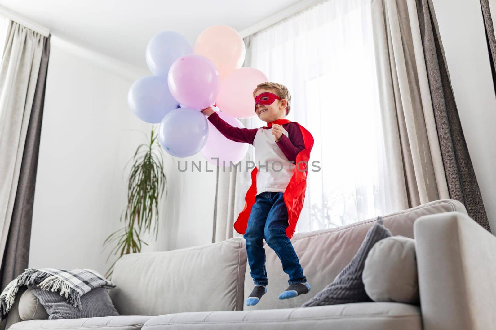 Happy boy in superhero costume playing with balloons, indoor fun. Dynamic kid portrait for invitation and greeting.