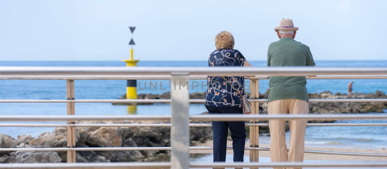 Captured in a tranquil moment, an elderly couple stands by the railing, overlooking the calm sea. The maritime beacon stands as a silent witness in the distance,
