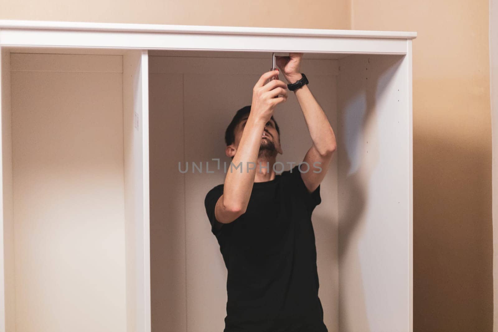 Young handsome caucasian brunette man is tightening screws with a screwdriver inside the wardrobe in the bedroom, closeup side view with selective focus. Furniture assembly concept.