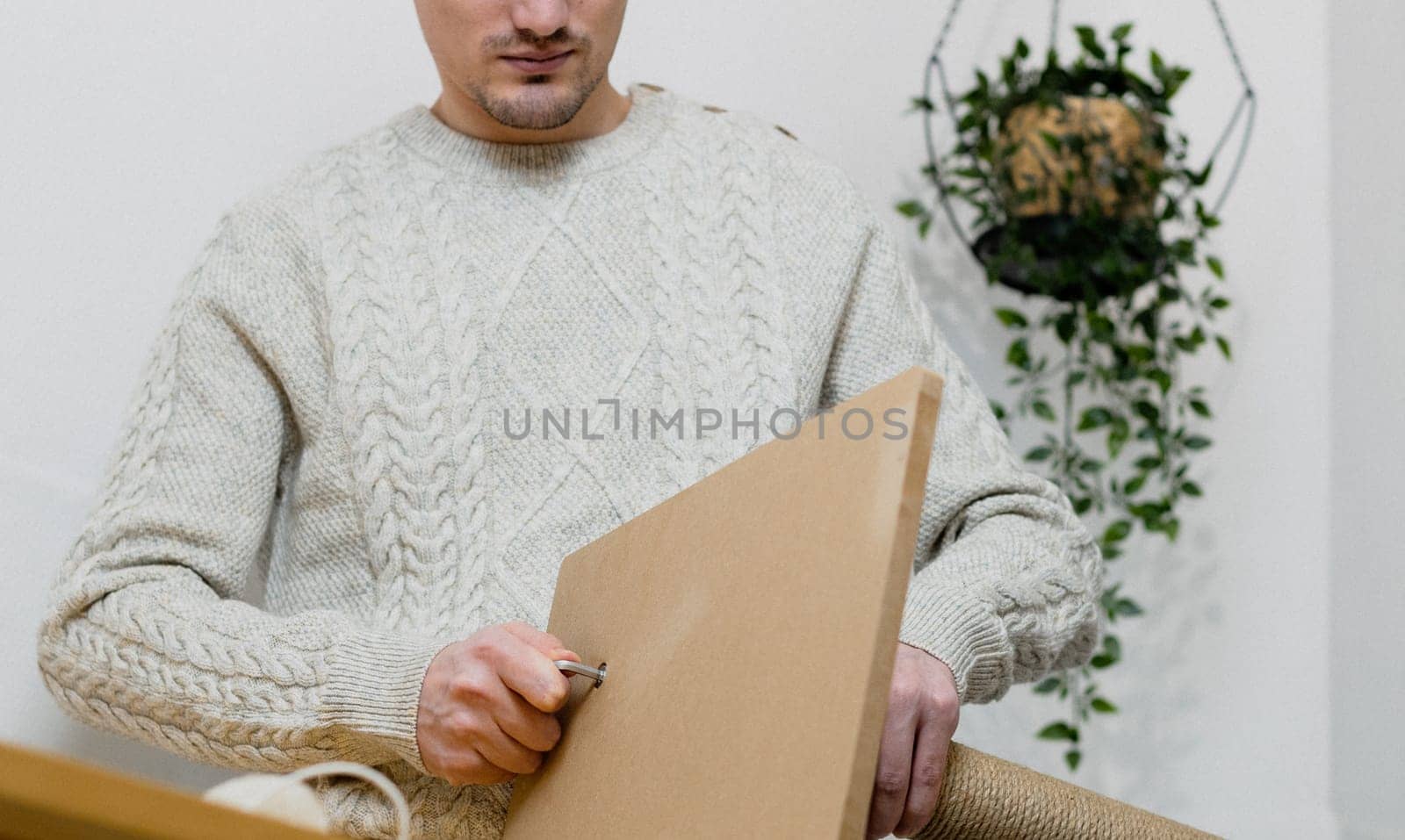 Young caucasian man in a knitted sweater and jeans manually drives a screw into a board while assembling a cat scratching post, standing at a table against a white wall with a hanging flower, close-up view from below. Concept assembly of a scratching post, cat furniture.
