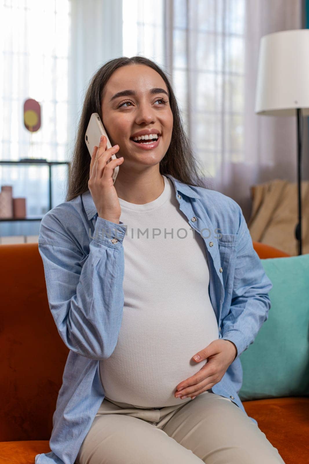 Smiling pregnant woman talking on smartphone sitting on sofa in living room at home. Happy lady stroking belly holding cellphone in apartment. Maternity prenatal care and pregnancy concept. Vertical