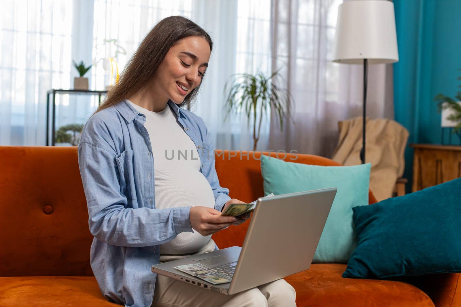 Happy pregnant woman counting money pressing keyboard keys on laptop smiling. Positive beautiful Caucasian expectant lady sitting on sofa couch buying online at home indoors. Comfort and convenience.