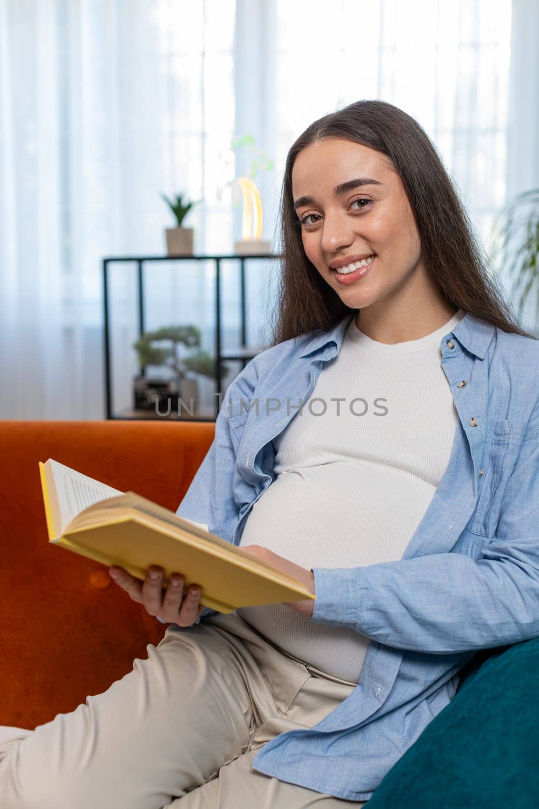 Pregnant Caucasian woman lady reading book feeling relax on cozy sofa couch at home. Future mother beautiful female girl read advice child care guide prepare prenatal. Relaxation and pregnancy concept