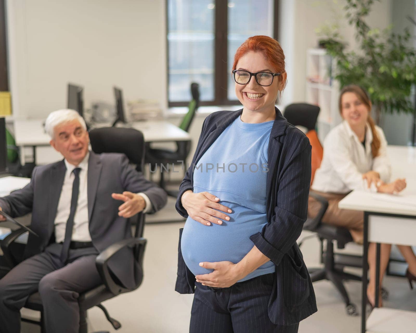 A happy pregnant woman stands in the middle of the office next to a Caucasian woman and an elderly man working at computers. by mrwed54