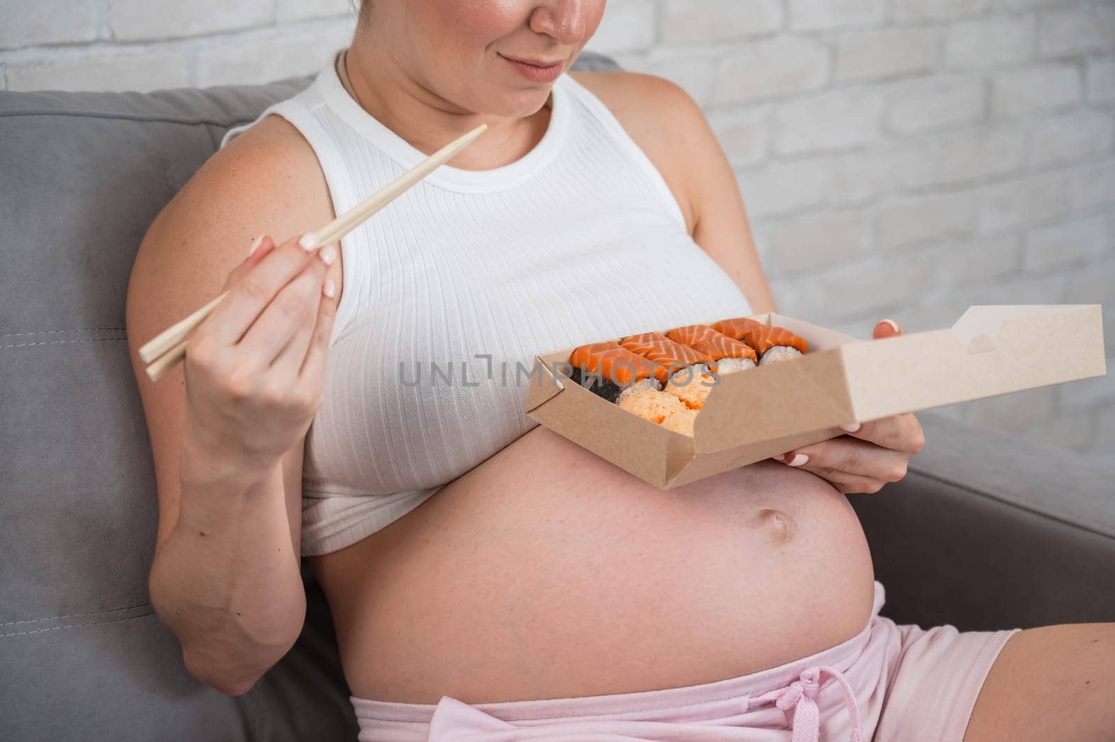 A pregnant woman sits on the sofa and eats rolls from a box. Food delivery. by mrwed54