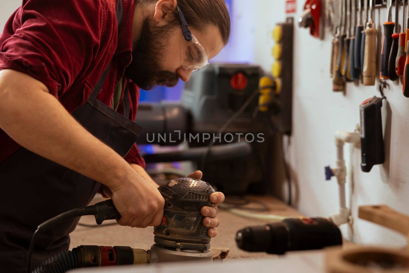 Carpenter wears protective glasses at workbench, using orbital sander with fine sandpaper on lumber to achieve refined finish. Man in assembly shop uses angle grinder on wood for professional results