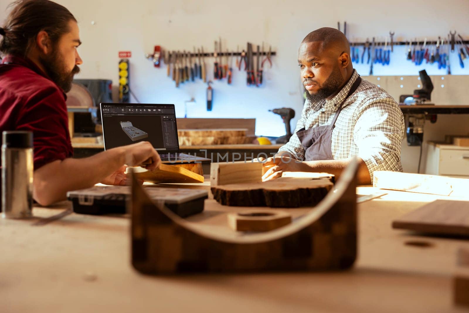 Cabinetmaker and colleague comparing wood piece used for furniture assembly with schematic displayed on laptop. Carpenter team in joinery crosschecking wooden object with notebook blueprint