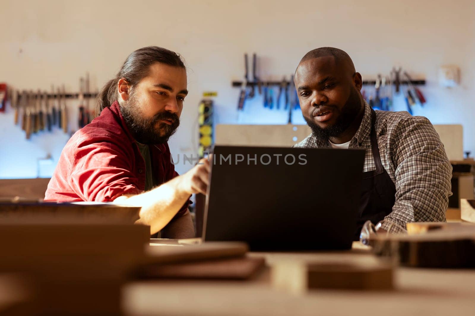 Woodworker looking over blueprints on laptop, brainstorming with colleague next steps in wood processing. Manufacturer and BIPOC apprentice reading furniture assembly schematics in studio