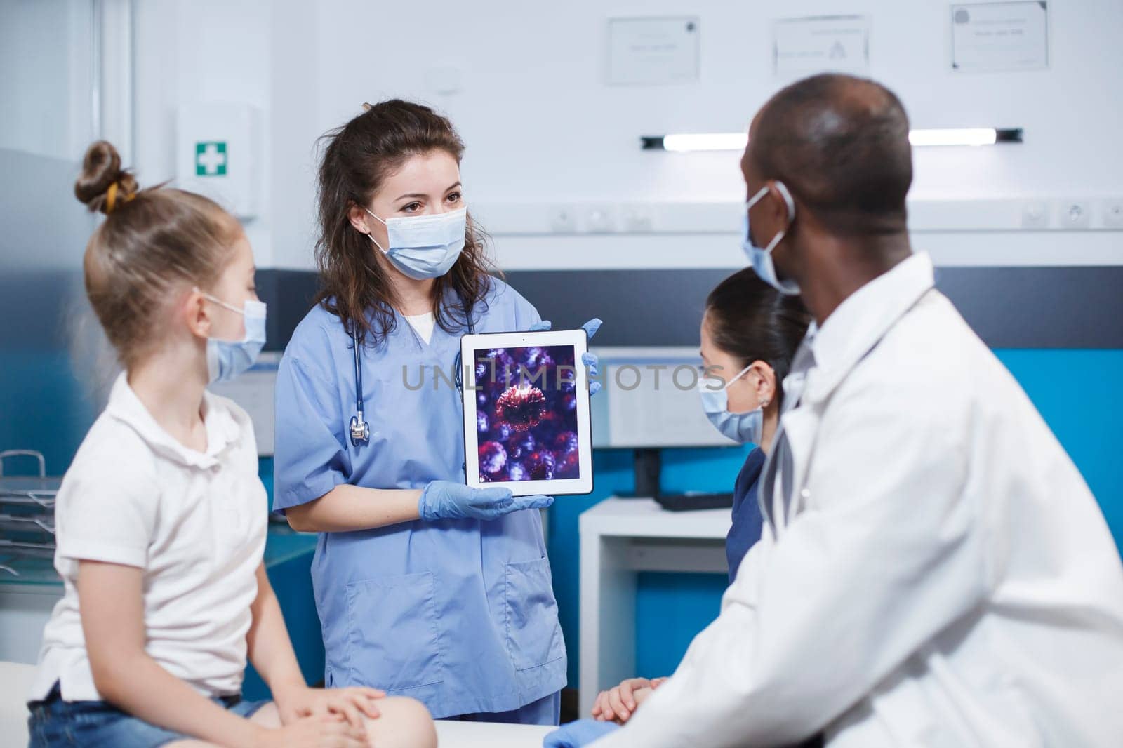 Woman wearing blue scrubs holding a digital tablet presenting a virus cell picture in front of a girl and her mother, and the african american male physician. Expert treatment and education provided.