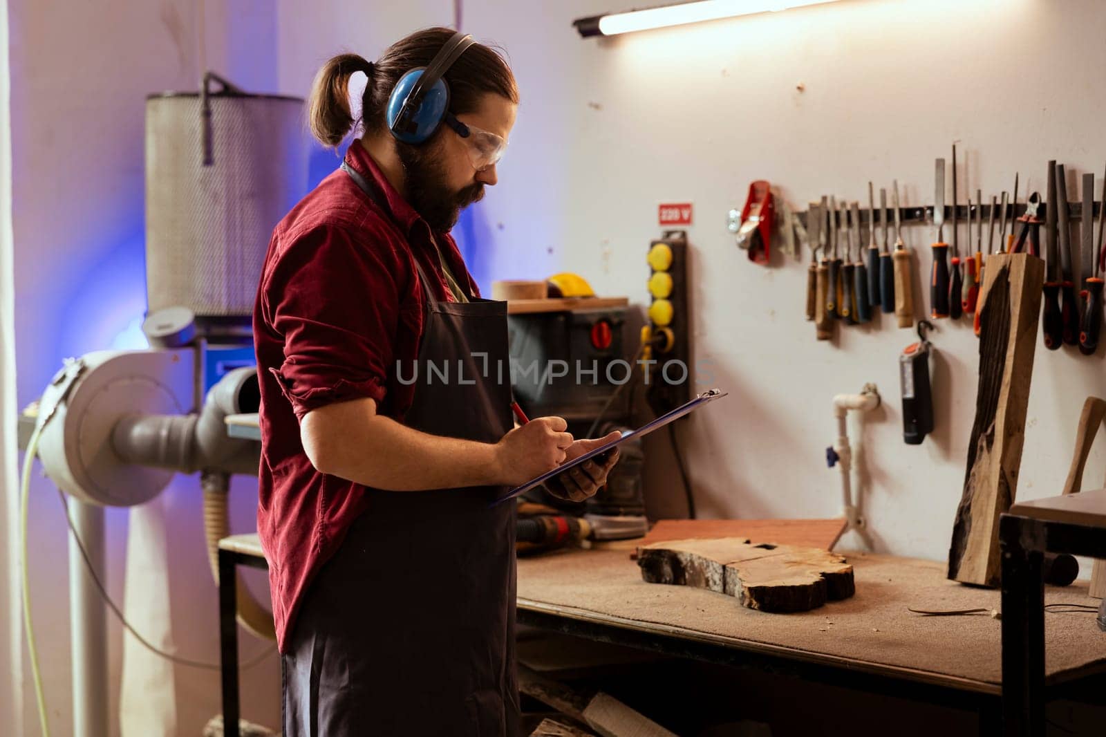 Carpenter wearing safety glasses drawing blueprints on notepad to make creative wood art pieces. Artisan looking at technical schematics to execute woodworking projects, using protective gear