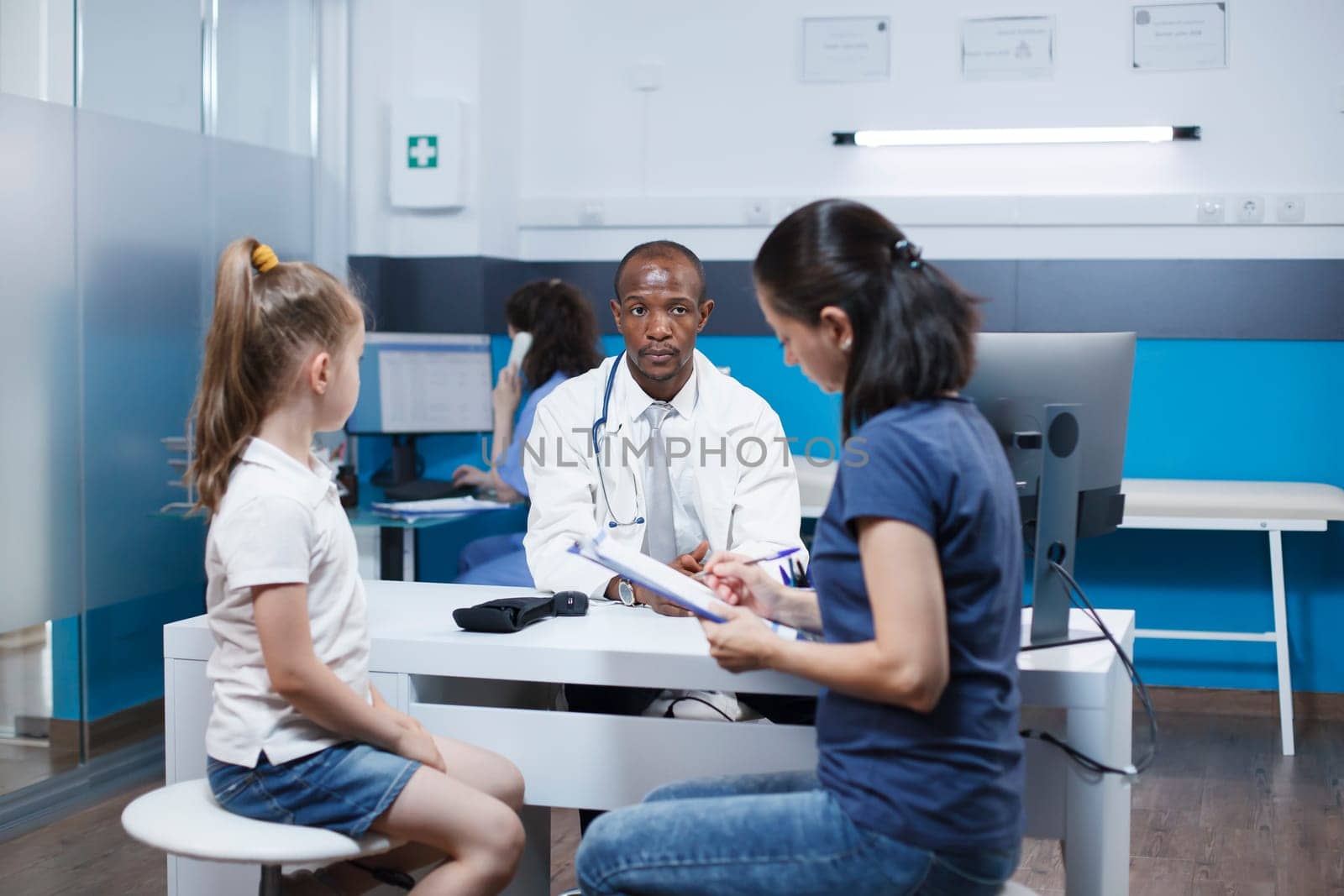 Filling out paperwork for a medical exam at the hospital office with her kid is a Caucasian mother. While the woman fills out papers at the clinic an African American doctor waits.