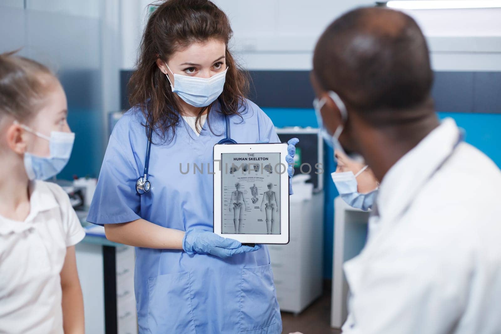 During a visit, bone doctor and nurse discuss the best medical alternatives for the patient in the clinic office. A doctor in antibacterial protective gear displays orthopedic information on a tablet.