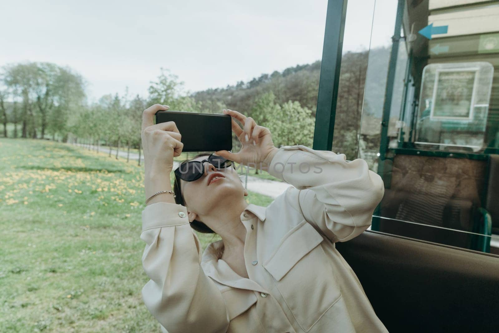 One beautiful young Caucasian brunette girl in sunglasses and stylish clothes takes pictures of nature on a mobile phone looking out of an open carriage of a tourist train on a spring sunny day in a public park, close-up side view.