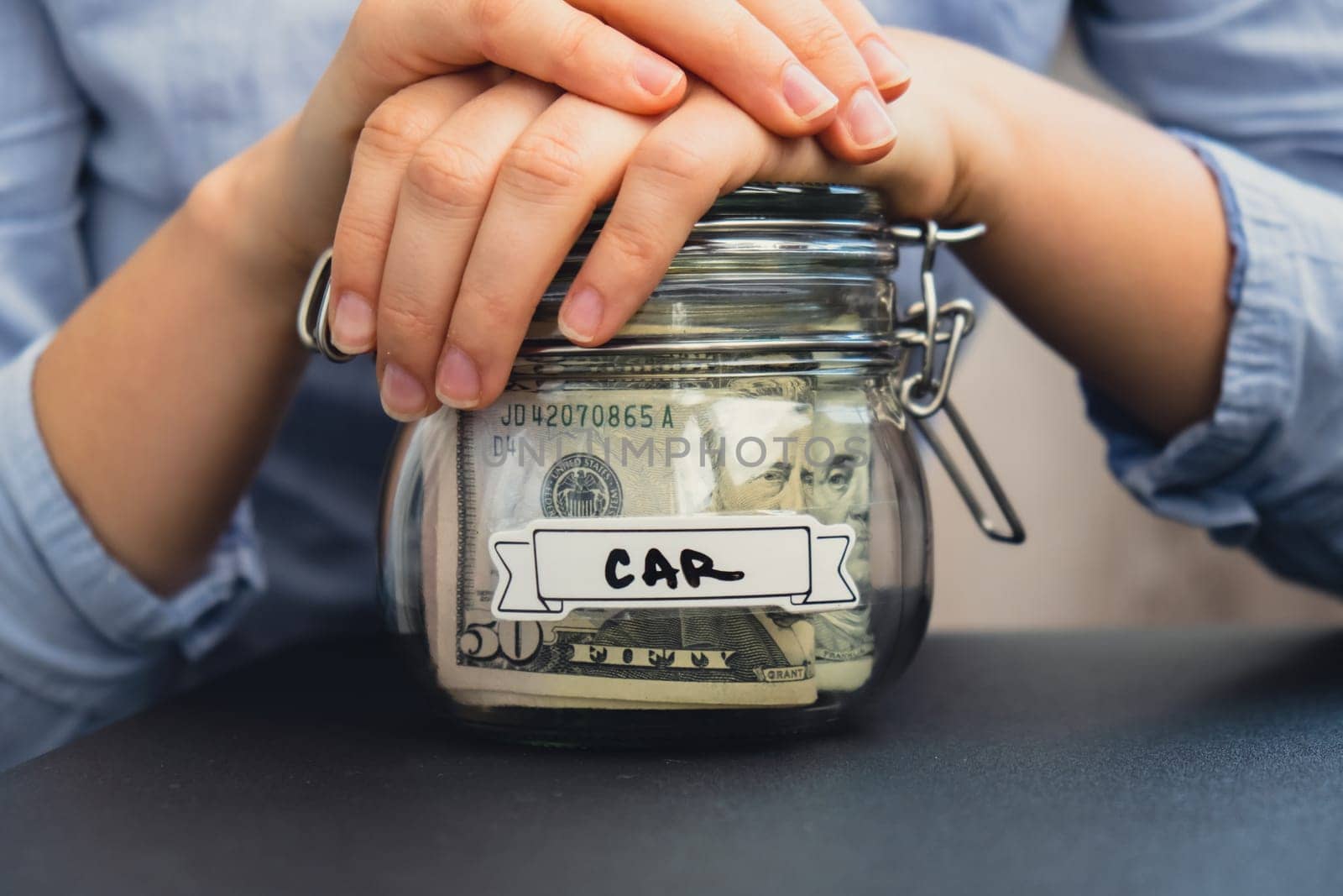 Unrecognizable woman holding Saving Money In Glass Jar filled with Dollars banknotes. CAR transcription in front of jar. Managing personal finances extra income for future insecurity by anna_stasiia