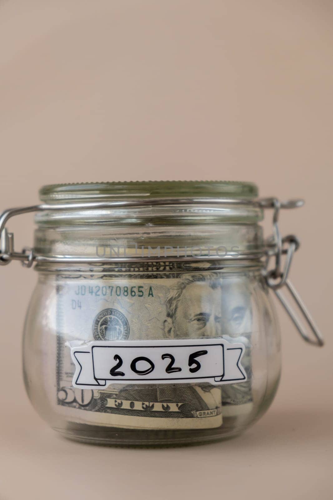 Saving Money In Glass Jar filled with Dollars banknotes. 2025 year transcription in front of jar. Managing personal finances extra income for future insecurity. Beige background