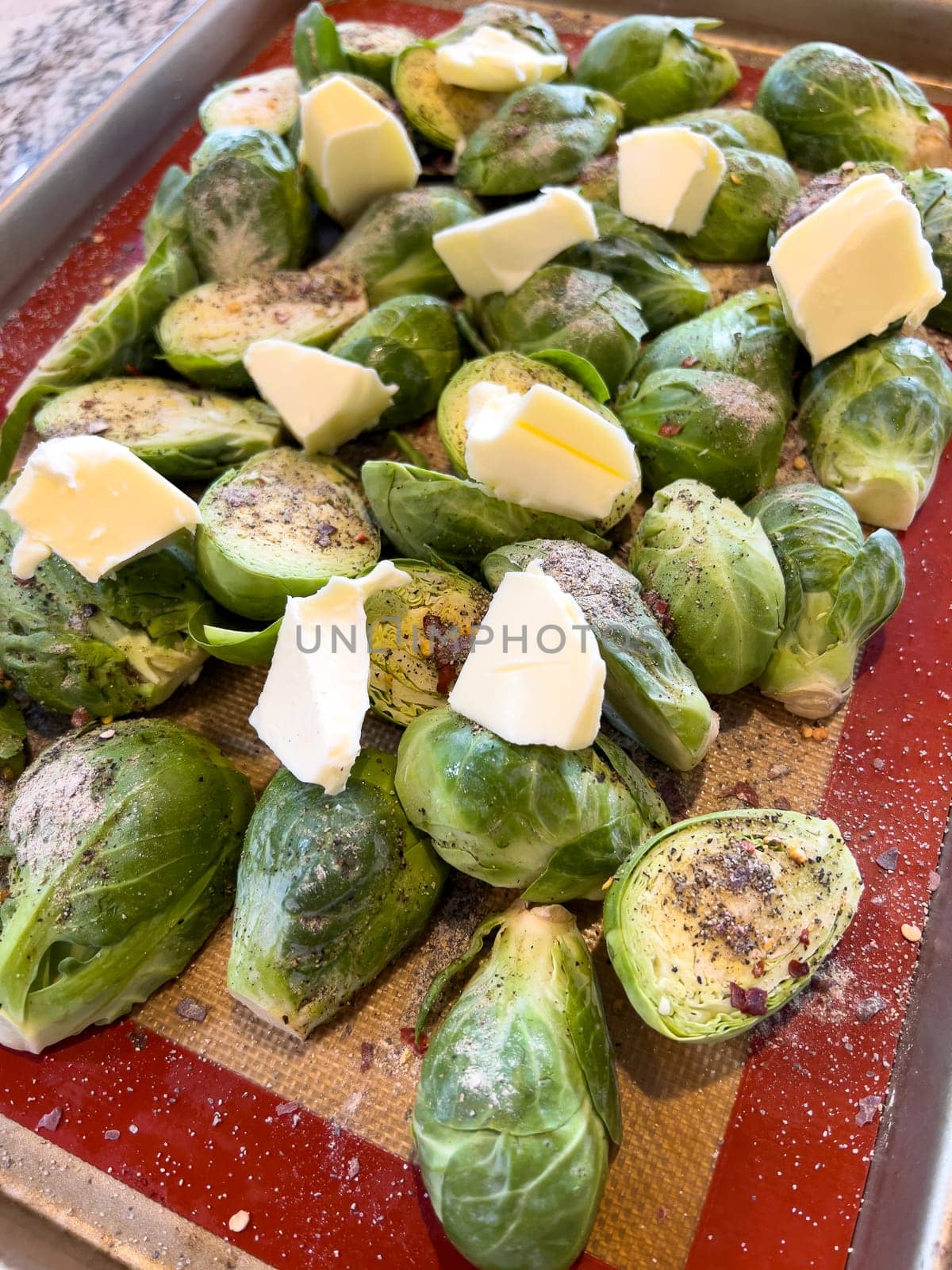 A tray of whole Brussels sprouts is generously topped with butter pats and seasoned, set against a non-stick baking sheet, indicating preparation for a savory roast.