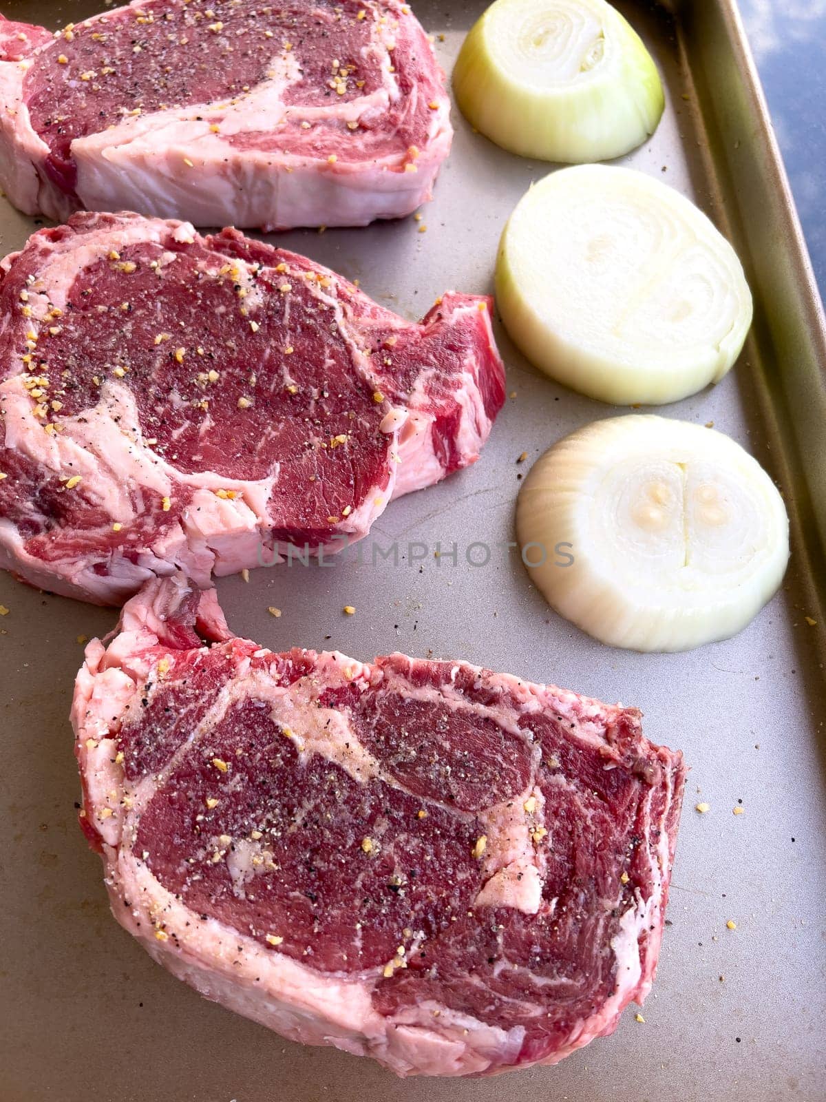 Three raw ribeye steaks with marbling, liberally seasoned with coarse spices, lie beside cut halves of onions on a kitchen tray, ready for cooking.