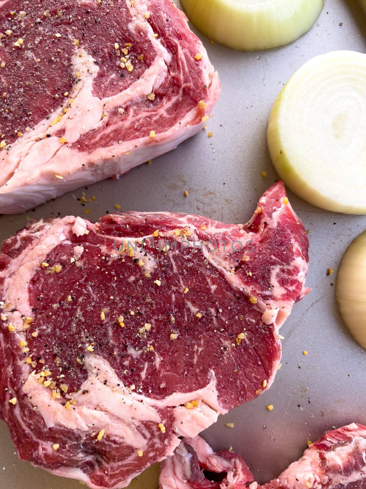 Raw Ribeye Steaks Seasoned with Spices and Onion Halves on Tray by arinahabich