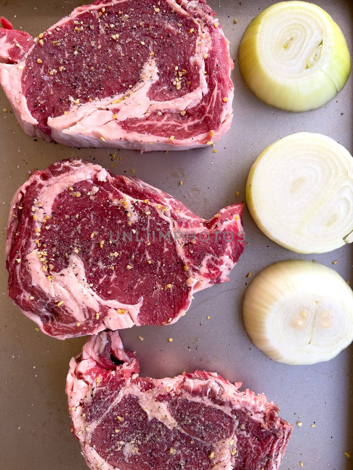 Three raw ribeye steaks with marbling, liberally seasoned with coarse spices, lie beside cut halves of onions on a kitchen tray, ready for cooking.